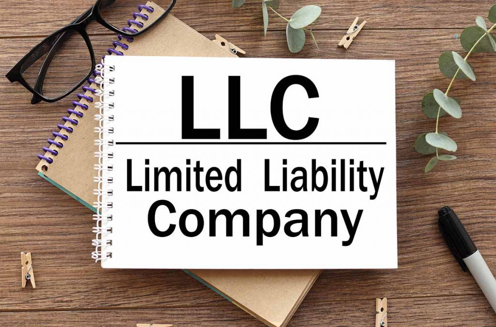 wooden desk with glasses, notepads and pens on it with a notepad that states LLC, Limited Liability Company.